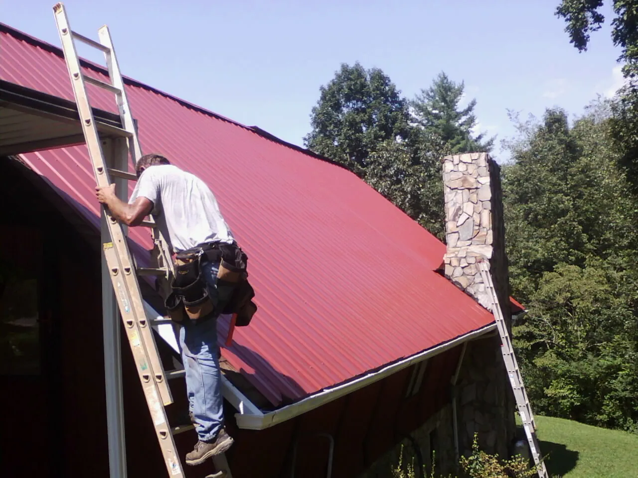 Roofer on a ladder beside a red metal roof and stone chimney — Soffit & Fascia Repair by Roofing Brothers in Womelsdorf